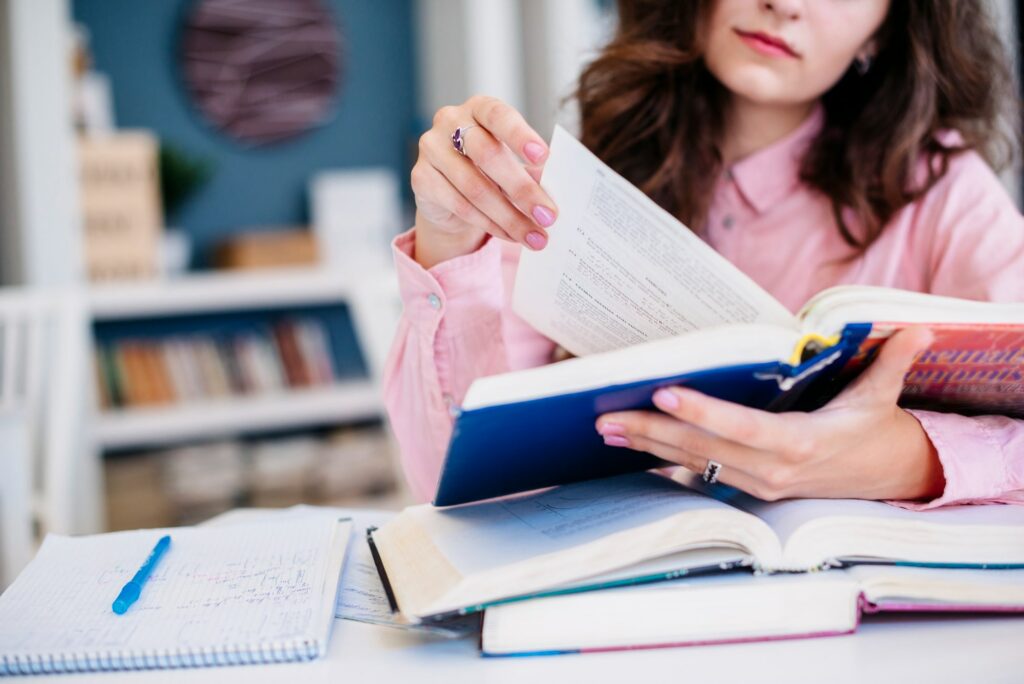 Jeune femme en train de lire un livre dans une bibliothèque. Sur le bureau, d'autres livres, un carnet et un stylo. 