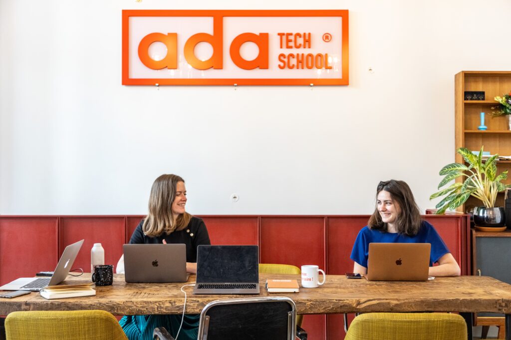 Deux femmes assises devant leurs ordinateurs se sourient à Ada Tech School. 