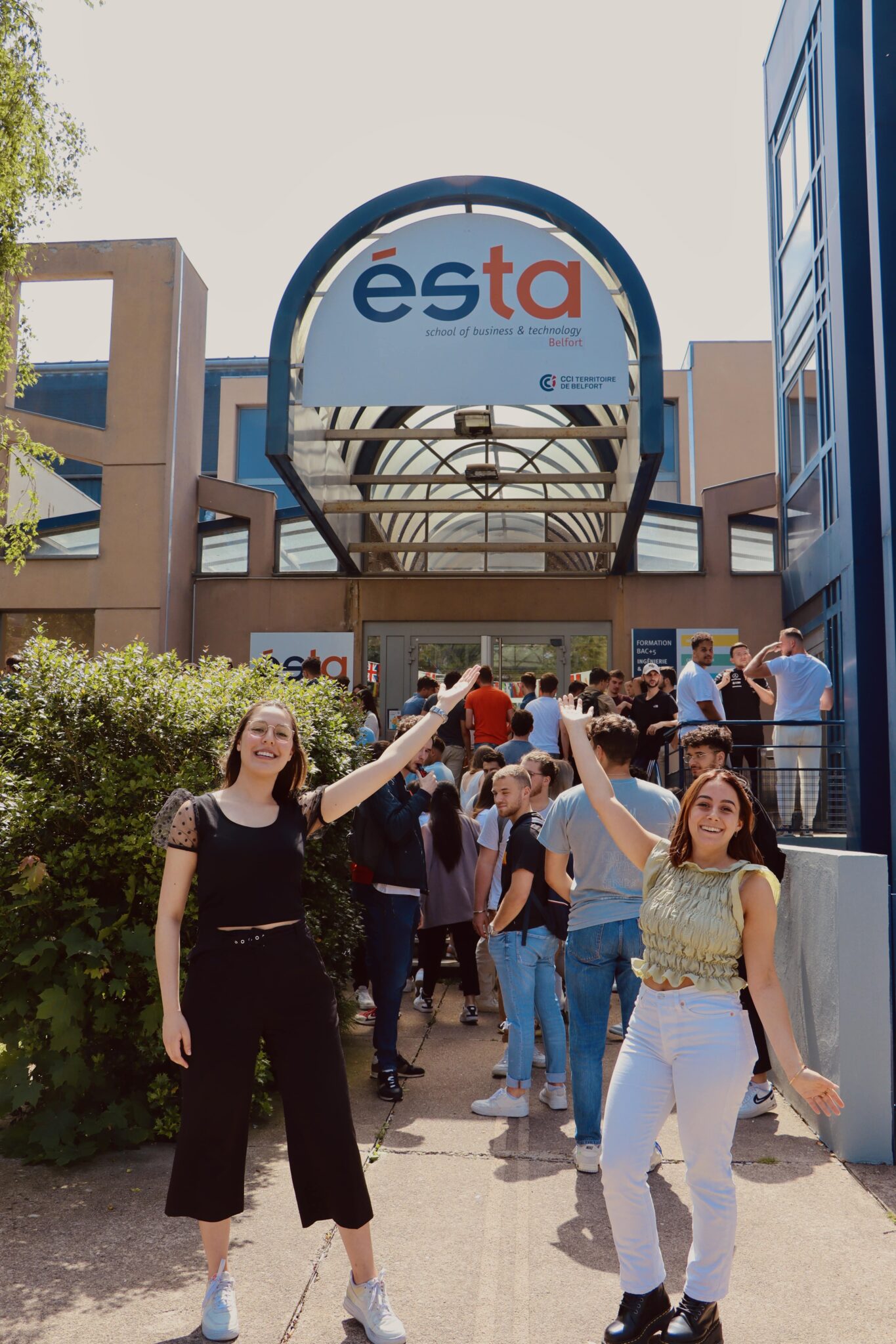 Deux jeunes étudiantes se tiennent devant l'ÉSTA, école pour devenir ingénieur commercial, et la présentent, sourires aux lèvres. 