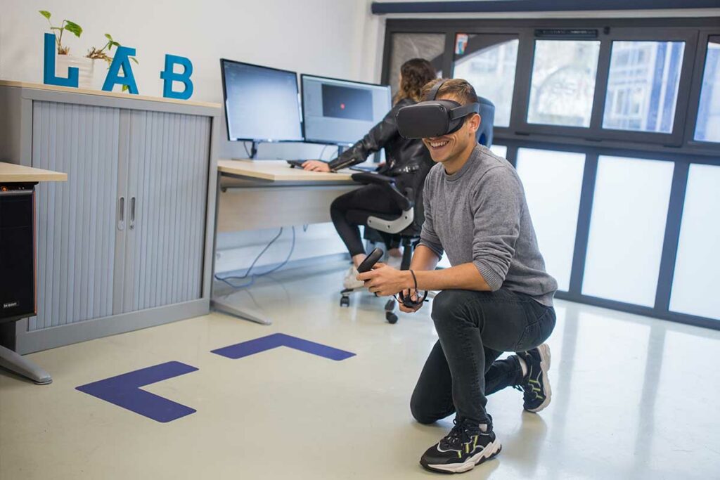 Jeune homme souriant au milieu du Digital Lab de l'ÉSTA, école pour devenir ingénieur commercial, portant un masque de réalité virtuelle sur les yeux. 